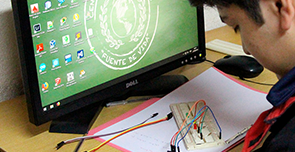 Fotografía de un joven estudiante frente a una computadora y un tablero didáctico de instalaciones eléctricas